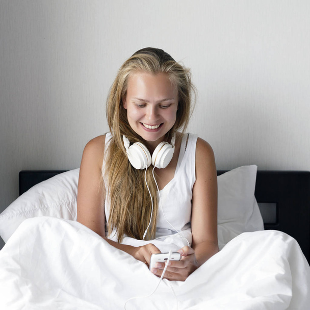 Young blond and happy smiling woman listen music and drink coffee in white bed in morning time. - Foto, immagini