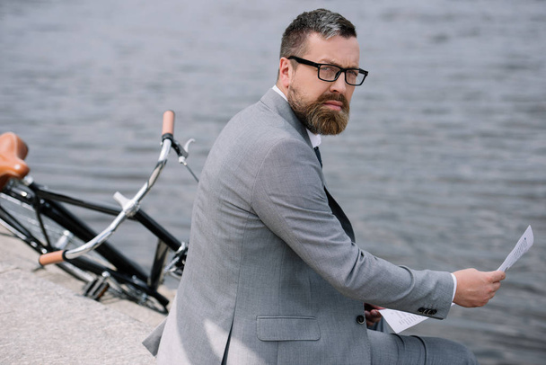 bearded businessman with newspaper sitting on quay with bike - Foto, Imagen