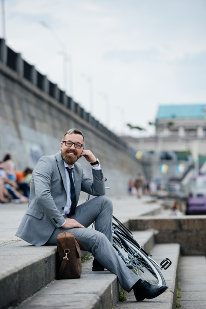 Bisiklet ile merdivenlerde oturan gri elbiseli gülümseyen iş adamı  - Fotoğraf, Görsel