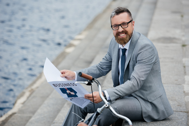 guapo hombre de negocios sonriente en traje gris leyendo periódico en muelle con bicicleta
 - Foto, imagen