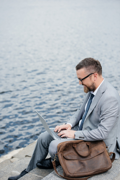 podnikatel, pomocí přenosného počítače a sedí na nábřeží s kožené pouzdro  - Fotografie, Obrázek