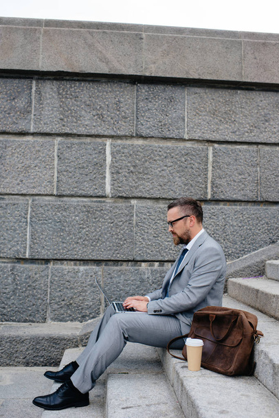 bearded businessman with bag and disposable cup of coffee using laptop and sitting on stairs - Photo, Image