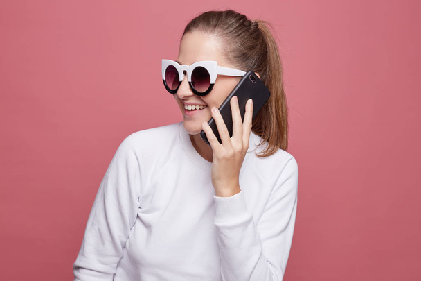 Studio profile shot of inspired woman laughs happily, has mobile conversation with close friend, enjoys leisure, poses on pink , uses modern electronic gadget. Fashionable lady in trendy spectacles. - Photo, image
