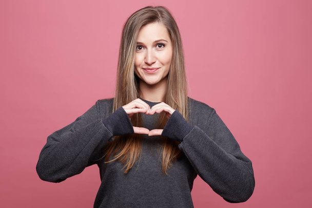 Pleased lovely Caucasian female with friendly look, keeps hands on heart gesture, demonstrates her good attitude, dressed in casual long sleeved shirt, expresses sincere feelings, love and compassion. - Photo, Image