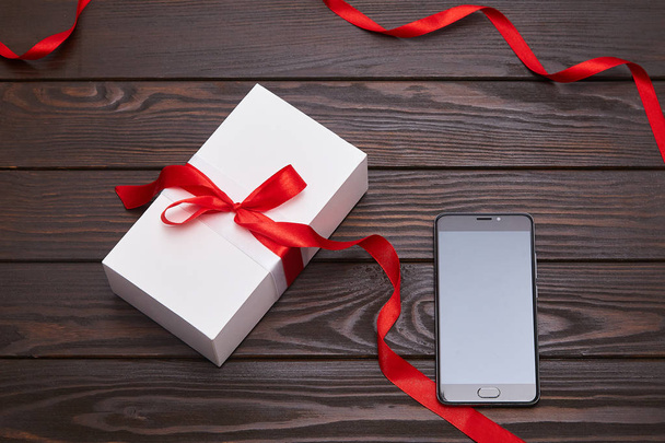 Białe pudełko z czerwoną wstążką i smartfon na ciemnym drewnianym tle.  - Zdjęcie, obraz