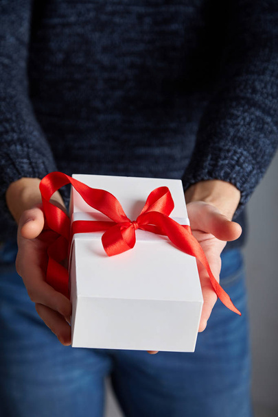  Белая подарочная коробка с красной лентой в мужских руках. Мягкий фокус
 - Фото, изображение