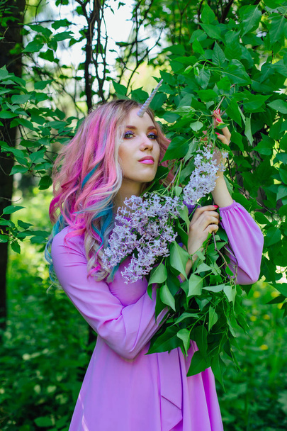 Fairy girl unicorn with rainbow hair and shiny makeup holding lilac flowers. - Photo, Image
