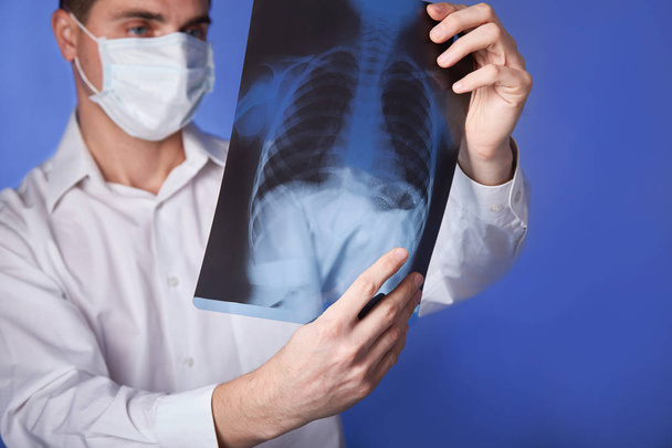 Lekarz w maski i biały płaszcz, trzymając RTG lub rentgen płuc, fluorography, obrazu na niebieskim tle - Zdjęcie, obraz