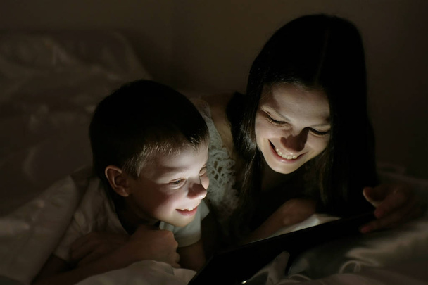 Young woman mom and her son watching a funny film together on tablet under the blanket. - Valokuva, kuva