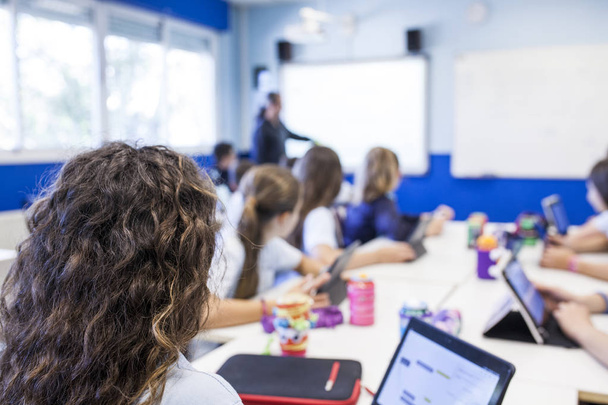 chica con pelo castaño rizado estudia en clase con su tableta al lado de sus compañeros de clase mientras escuchan a la maestra
 - Foto, Imagen