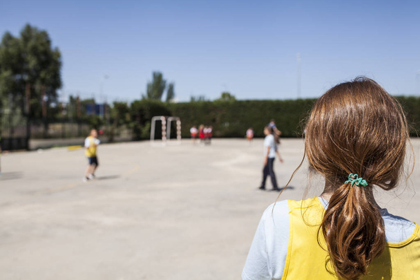 руда дівчина з жовтим жилетом дивиться, як її однокласники грають на уроці фізичного виховання на ігровому майданчику в школі в сонячний день
 - Фото, зображення