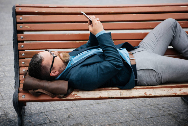 businessman using smartphone lying on bench in city - Photo, Image