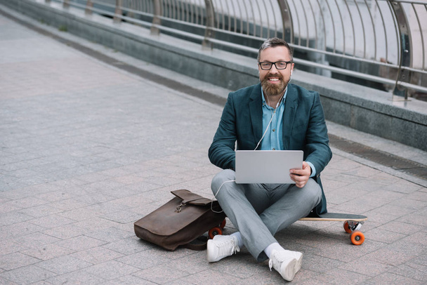 stylish bearded man using laptop and sitting on longboard with leather bag in city - Zdjęcie, obraz