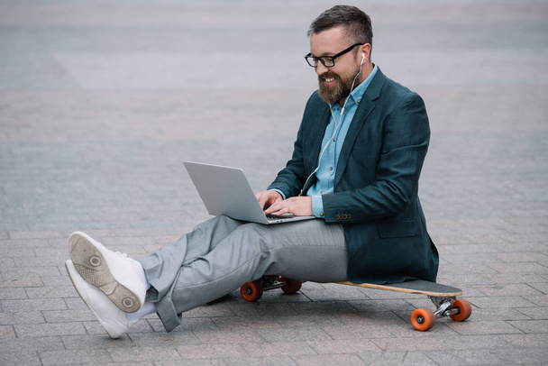 stylish european man using laptop and sitting on skateboard in city - Foto, afbeelding