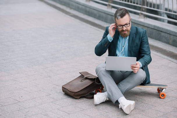 bearded man using laptop with earphones and sitting on skateboard in city - Foto, afbeelding