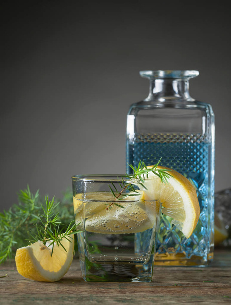 Голубой джин, тоник и лимонные ломтики на старом деревянном столе. Алкогольный коктейль с ломтиками лимона и можжевеловой веткой
. - Фото, изображение
