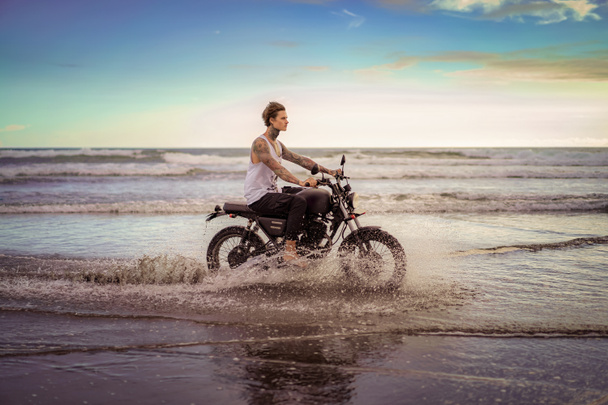 side view of handsome tattooed man riding motorcycle in ocean waves on beach - Photo, Image