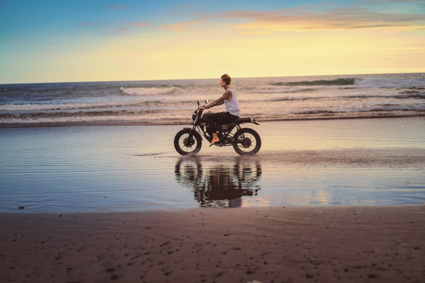 side view of tattooed biker riding motorcycle on ocean beach during sunrise - Photo, Image