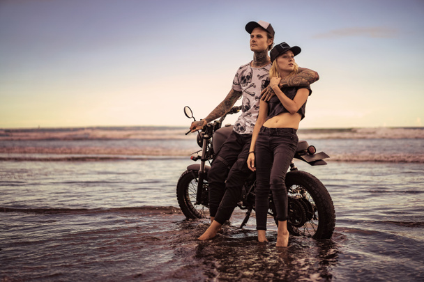 stijlvolle paar staande en knuffelen in de buurt van motorfiets op oceaan strand  - Foto, afbeelding