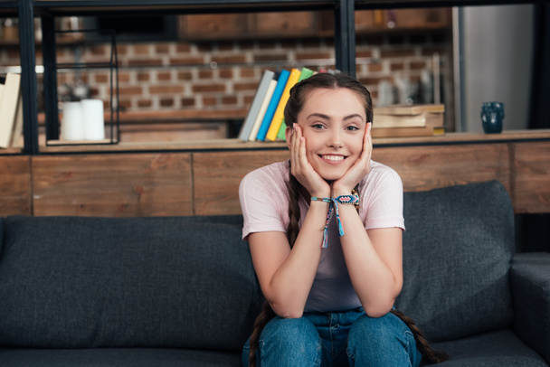 usmívající se dospívající dívka s rukama na tvářích při pohledu na fotoaparát a sedí na gauči doma - Fotografie, Obrázek