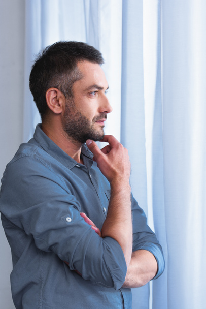 side view of thoughtful man with hand on chin in front of curtains at home  - Foto, afbeelding