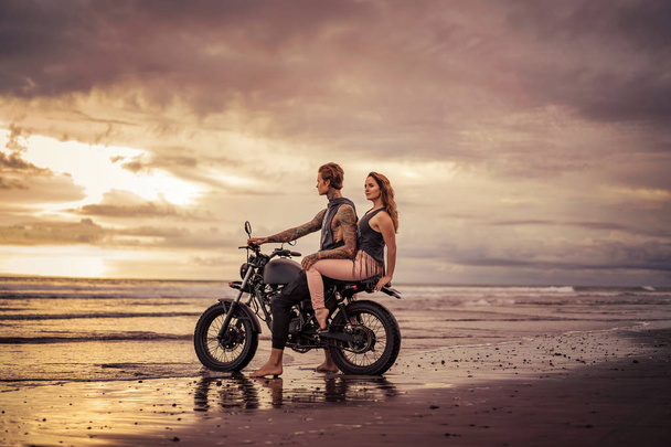 pareja cariñosa sentada en motocicleta en la playa durante el amanecer
  - Foto, Imagen