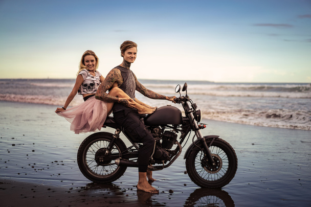 χαμογελώντας ζευγάρι αγκαλιάζει τη μοτοσικλέτα στην ωκεάνια παραλία κατά την όμορφη ανατολή του ηλίου  - Φωτογραφία, εικόνα