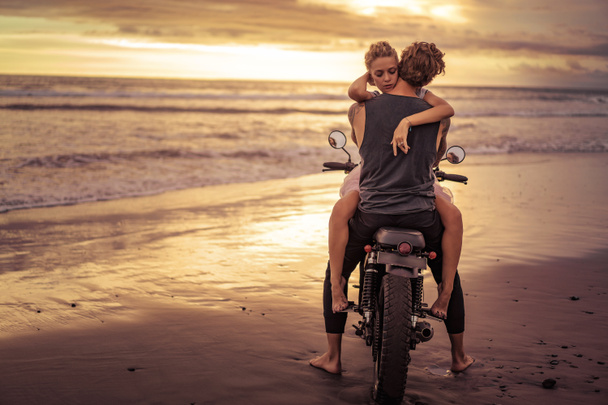 Heterosexuelles Paar umarmt sich auf Motorrad am Strand des Ozeans bei schönem Sonnenaufgang  - Foto, Bild