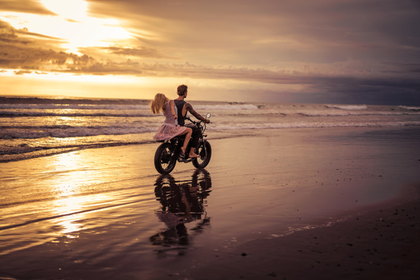 rear view of boyfriend and girlfriend riding motorbike on ocean beach during sunrise - Photo, Image
