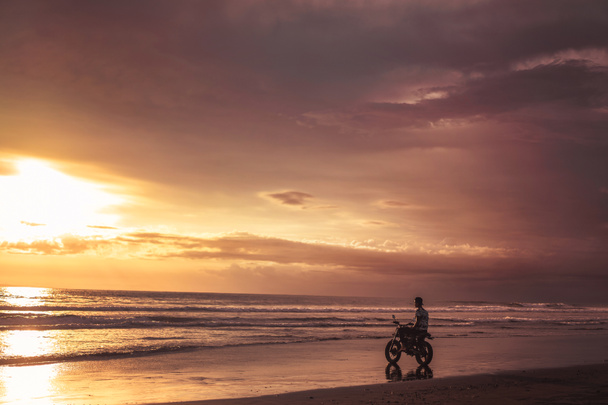 man sitting on motorbike on seashore and looking at beautiful sunset - Fotó, kép