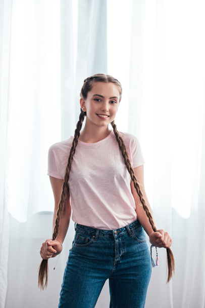 happy teenage girl looking at camera and holding own plaits in front of curtains at home  - Фото, зображення