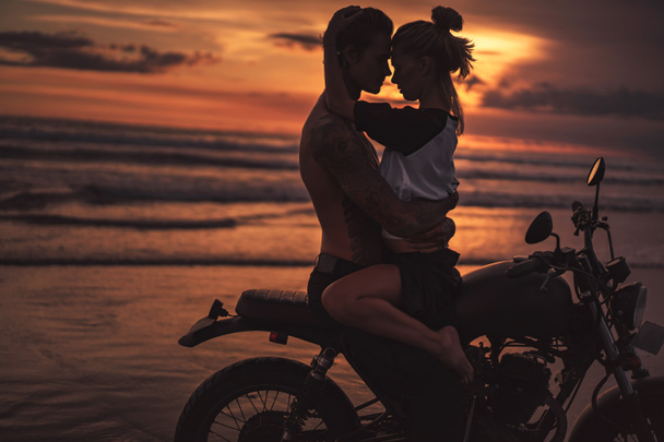 shirtless boyfriend hugging girlfriend on motorcycle at beach during sunset - Photo, Image