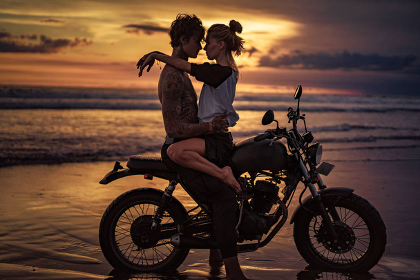 passionate couple cuddling on motorcycle at beach during sunset - Photo, Image