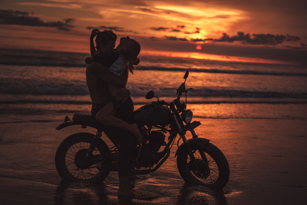 Σέξι ζευγάρι αγκαλιάζει την μοτοσυκλέτα στην παραλία κατά τη διάρκεια του ηλιοβασιλέματος - Φωτογραφία, εικόνα
