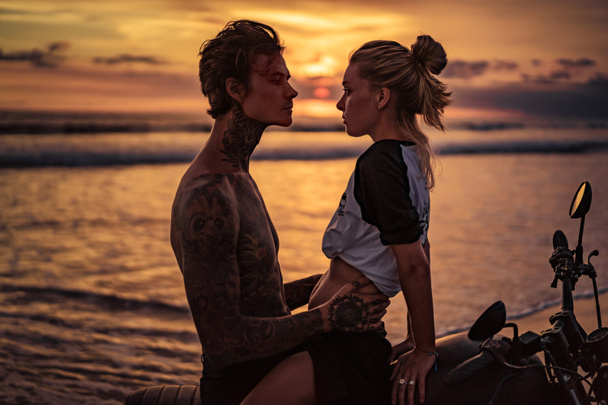 Παθιασμένος ζευγάρι αγκαλιάζει την μοτοσυκλέτα στην παραλία κατά τη διάρκεια του ηλιοβασιλέματος - Φωτογραφία, εικόνα