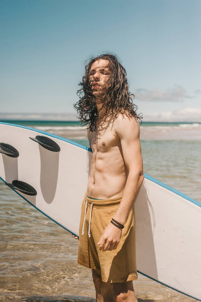 portrait of young man with surfboard standing in ocean on summer day - Фото, изображение