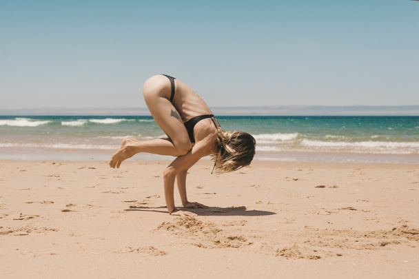 Seitenansicht einer jungen Frau im schwarzen Bikini, die am Sandstrand Bakasana-Yoga praktiziert - Foto, Bild