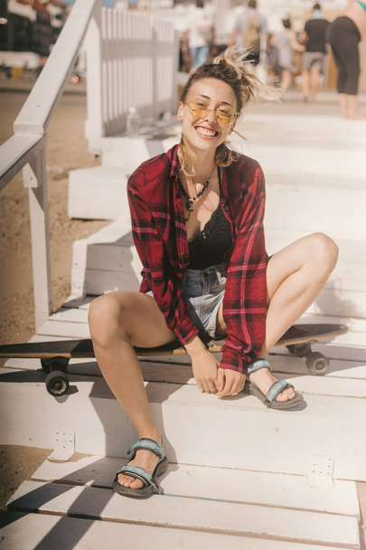 young smiling woman in sunglasses and shirt resting on longboard on wooden stairs on beach - Fotó, kép