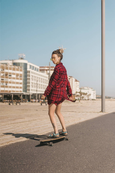 young woman in sunglasses and shirt skating on longboard on street in Portugal - 写真・画像