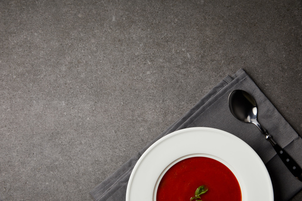 top view of tasty tomato soup in plate and spoon on grey napkin on grey table - Fotografie, Obrázek