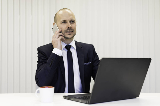 sonriente hombre de negocios con portátil sentado en la mesa mientras habla en el teléfono inteligente
  - Foto, Imagen