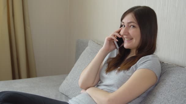 νεαρή γυναίκα κάθεται στον καναπέ και μιλάει στο τηλέφωνο - Πλάνα, βίντεο