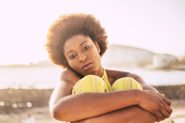 beautiful skin and face for nice black race African model with alternative afro hair. sad and lonely hugging herself at the beach with defocused ocean in background, summer alone leisure time. - Foto, Bild