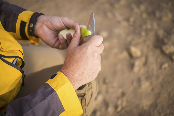 close up of human hands cutting a green apple with a knife outdoor during an adventure trekking outdoor in the desert. exploring the world and get an alternative travel and vacation concept - Photo, Image