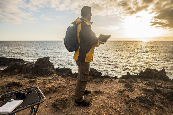 digitaler Nomadenreisender bei der Arbeit, der einen erstaunlichen goldenen Sonnenuntergang auf dem Ozean betrachtet. Kamera und Laptop und schönen Lebensstil in der Freizeit. Alternatives Arbeitsamt zum freien Leben - Foto, Bild