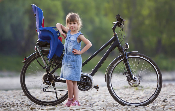 Petite jolie fille blonde en robe bleue debout sur des cailloux devant un vélo moderne avec siège enfant sur fond d'arbres verts flous le jour de l'été. Style de vie actif et concept de loisirs familiaux
. - Photo, image