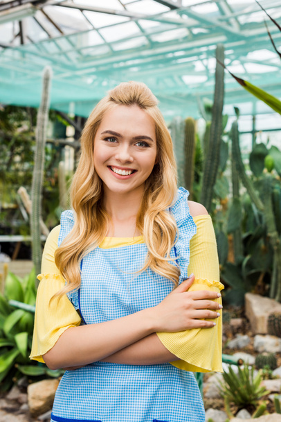 Krásná blondýna žena v zástěře stojí s překřížením rukou a usmívá se na kameru ve skleníku - Fotografie, Obrázek