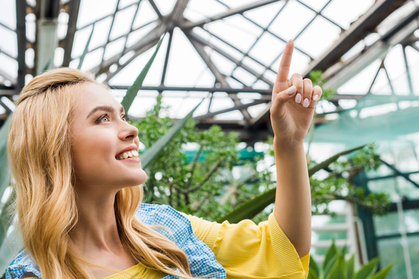 kaunis hymyilevä blondi nainen osoittaa ylös sormella kasvihuoneessa
 - Valokuva, kuva