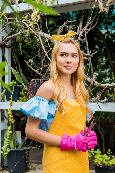 beautiful pensive blonde woman in apron and rubber gloves holding scissors and looking away in greenhouse - Photo, Image