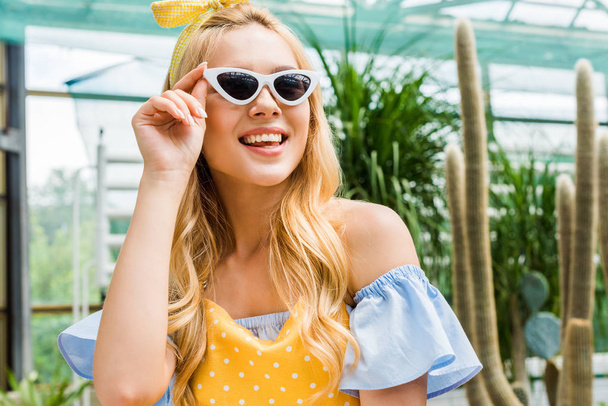beautiful happy blonde girl in sunglasses and apron smiling in greenhouse - Zdjęcie, obraz